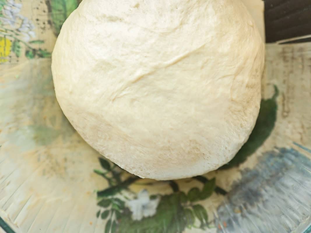 A ball of raw dough sits on a patterned glass plate. The dough is smooth and pale, resting atop a colorful floral design printed on the plate, which provides a contrasting backdrop.